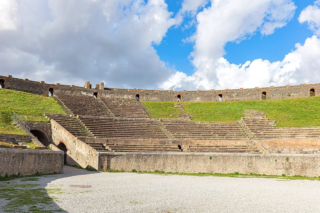 Die Arena und die grasbewachsenen Sitzreihen des Amphitheaters von Pompeji unter bewölktem blauem Himmel