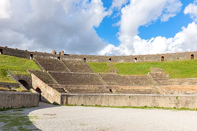 La arena y las gradas cubiertas de hierba del anfiteatro de Pompeya bajo un cielo azul con nubes
